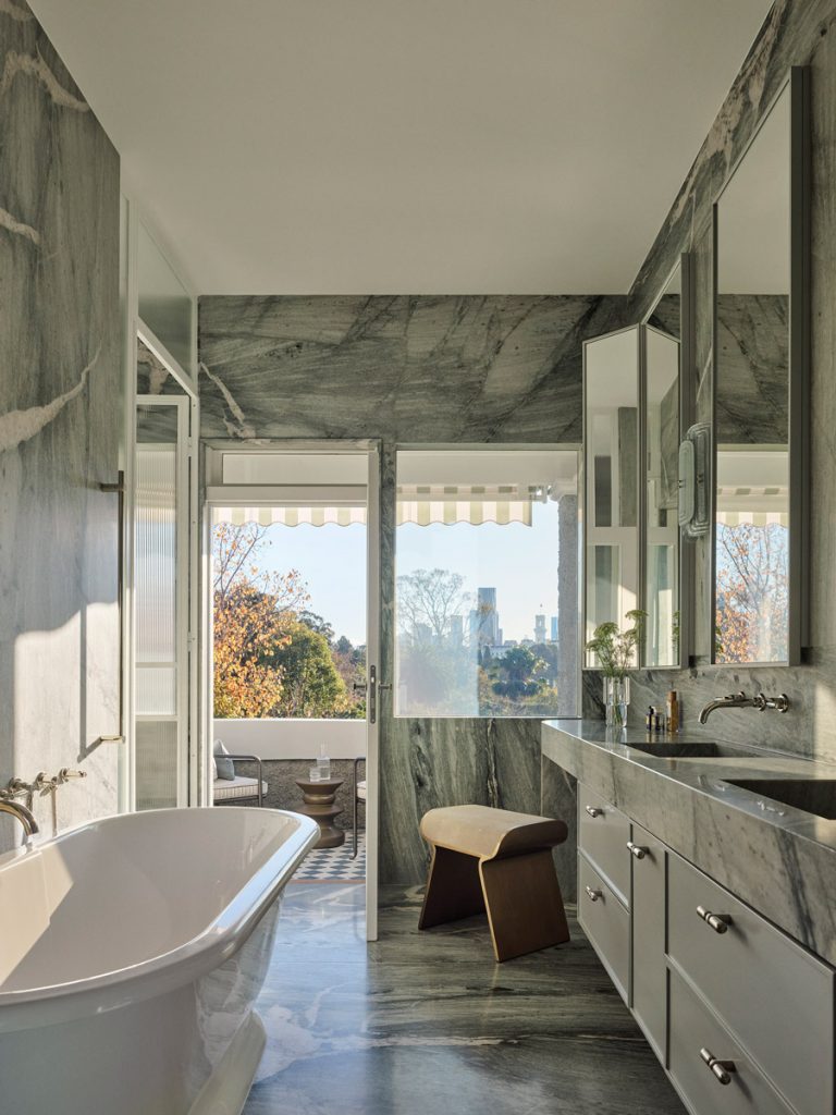 Luxurious bathroom with marble walls, a freestanding tub, sleek cabinetry, and a view through large windows framed by greenery.