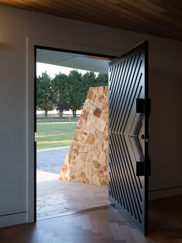 Bespoke black iron door with diagonal grooves, open to reveal a stone wall and green landscape outside, framed by a modern interior.