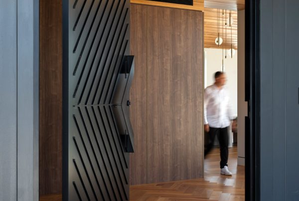 Bespoke black iron door with diagonal lines, opening to a warm wooden interior, featuring a blurred figure walking inside.