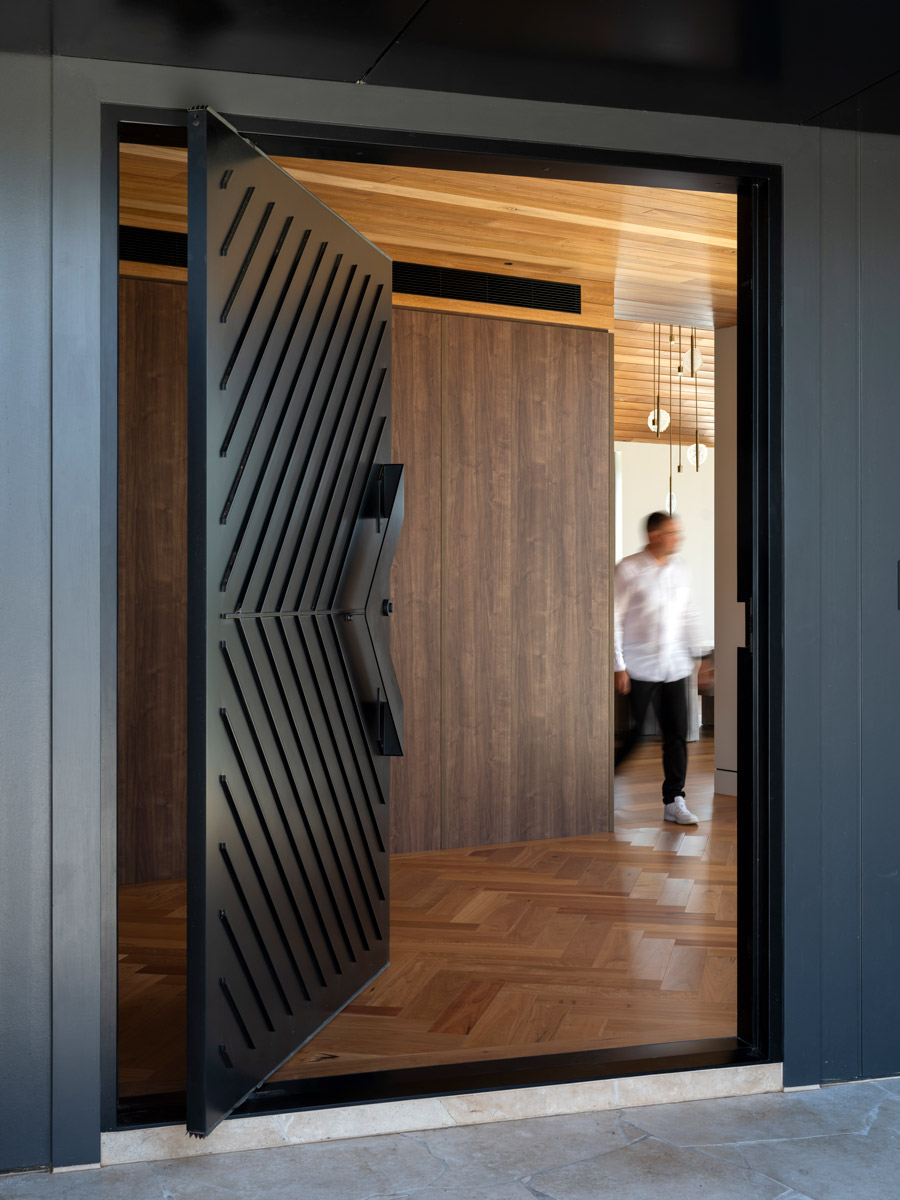 Bespoke black iron door with diagonal lines, opening to a warm wooden interior, featuring a blurred figure walking inside.